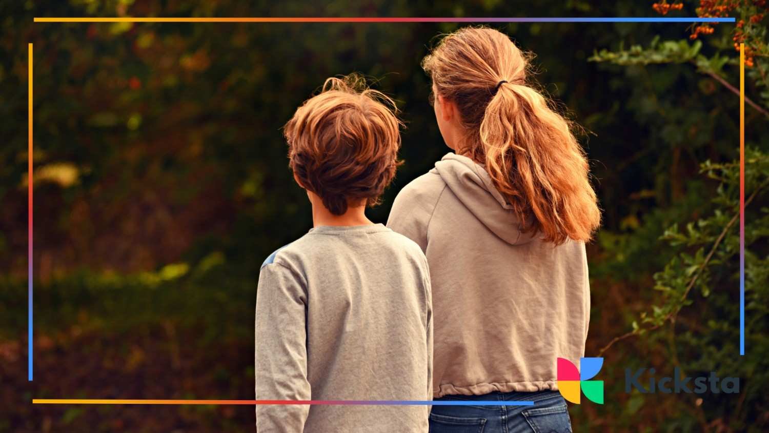 A girl and boy stand side by side, facing away, looking into a forest