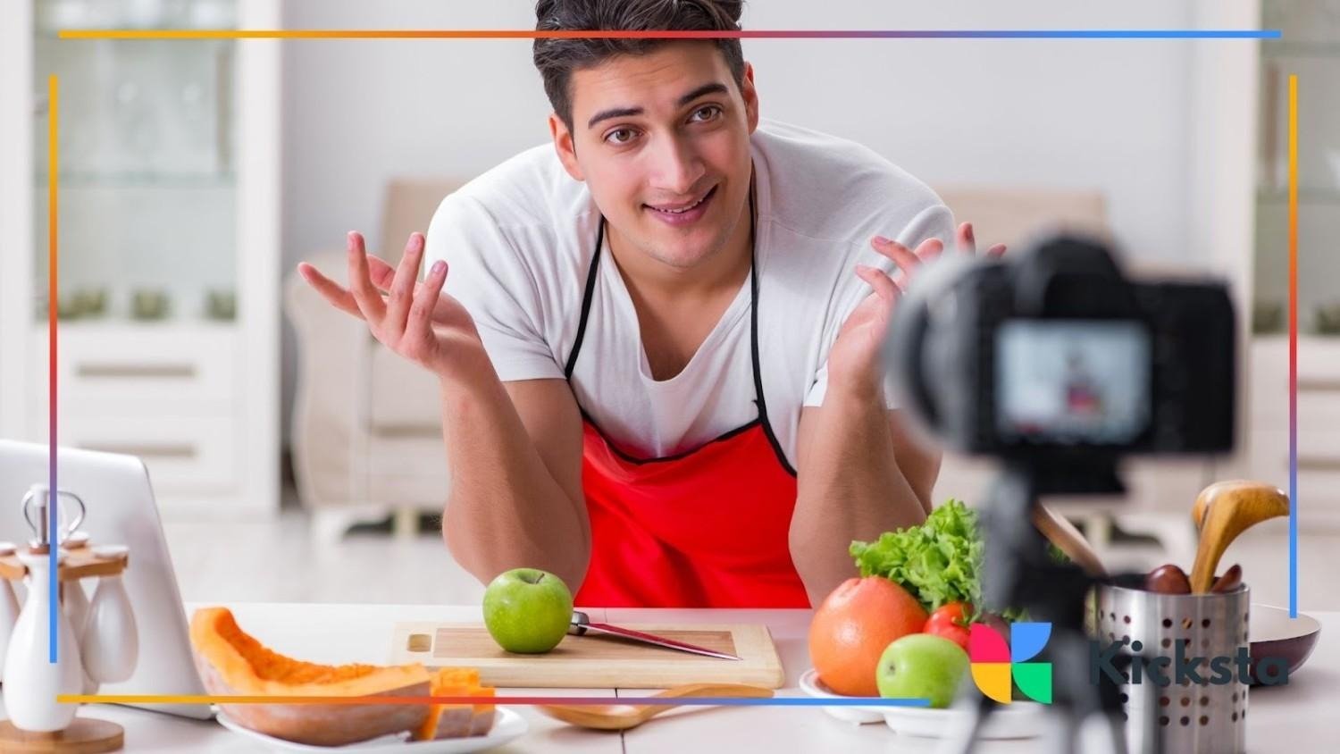 Man wearing a red apron recording a cooking video in the kitchen, surrounded by fruits and vegetables