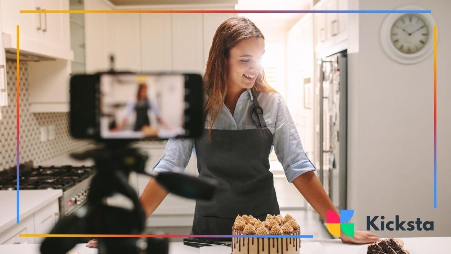 Woman in an apron smiling while filming a baking tutorial in the kitchen with cakes on the counter