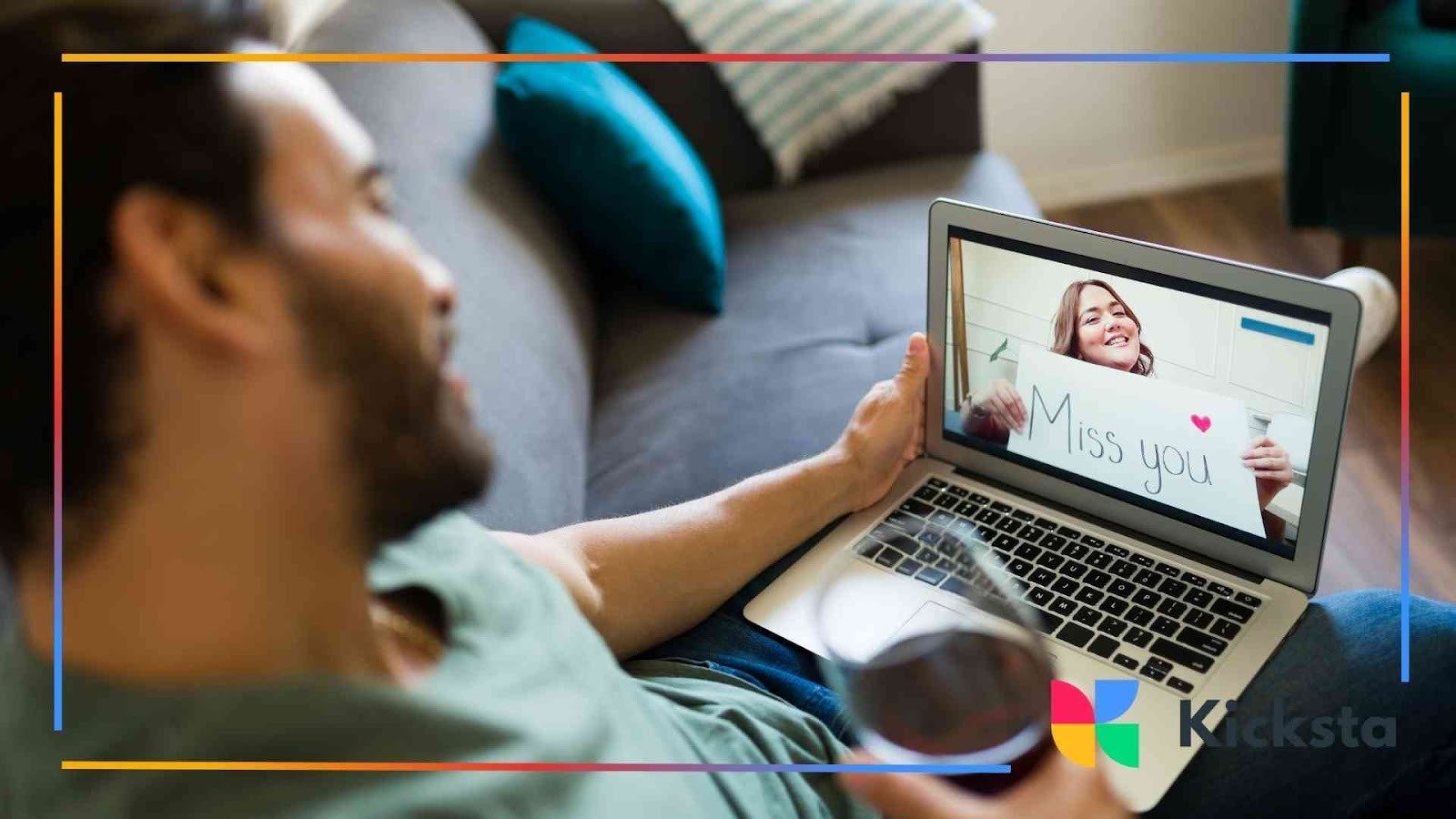 Man holding a glass of wine while smiling at his laptop screen showing a woman holding a sign that says “Miss you.”