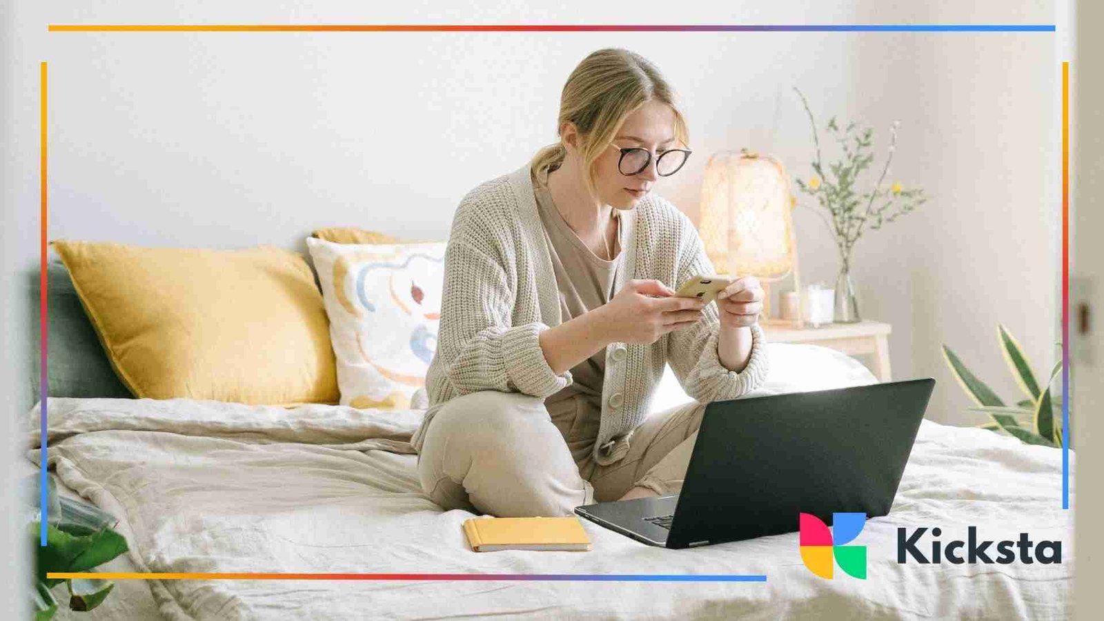 Woman sitting on her bed using her phone with an open laptop and notebook beside her.