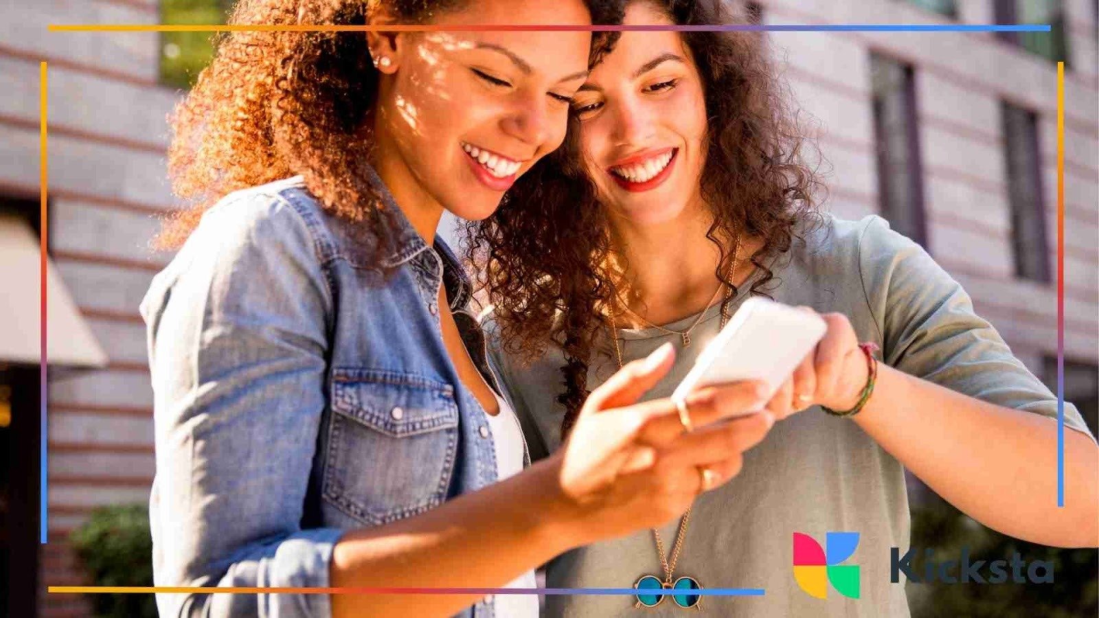 Two friends standing outdoors and smiling while looking at a smartphone together.