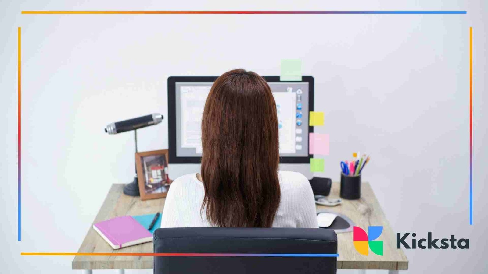A woman sits at a desk facing a computer monitor, surrounded by notebooks, sticky notes, pens, and a desk lamp in a clean, organized workspace.