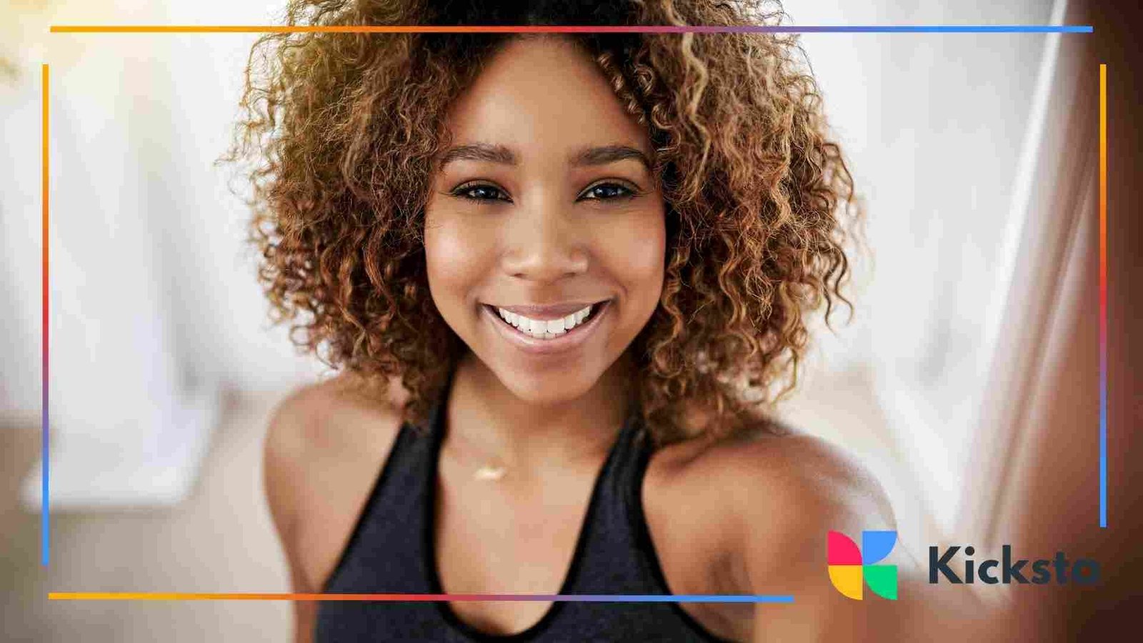 A close-up of a smiling woman with curly hair wearing a sleeveless workout top, standing indoors with soft natural light in the background.