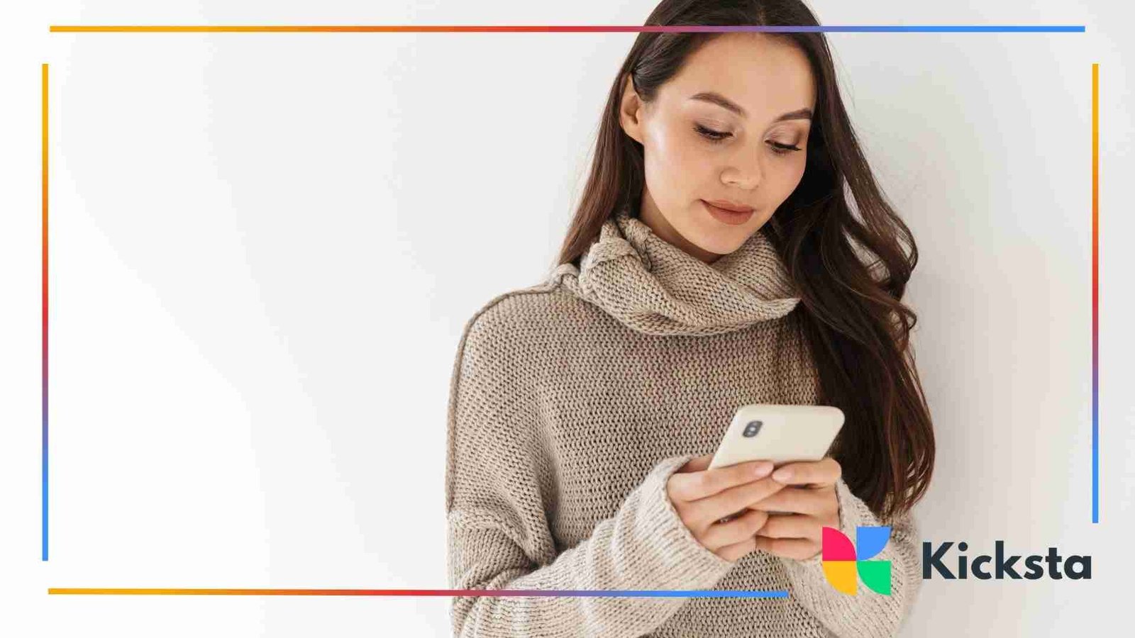 Young woman wearing a beige knitted sweater looking down at her smartphone while standing against a plain white background.