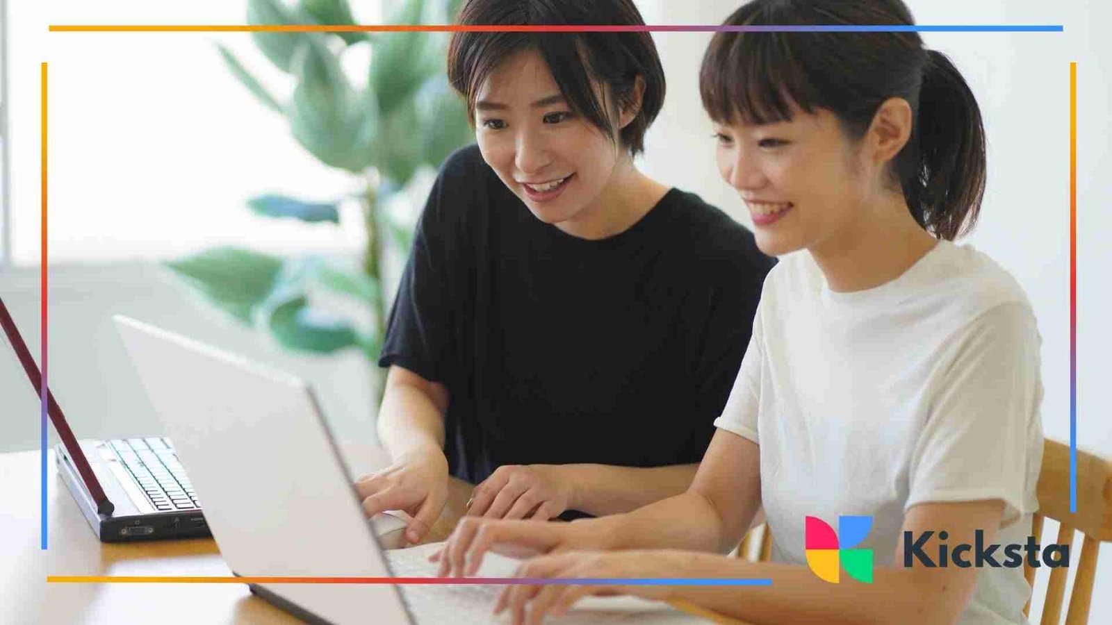 Two women sitting at a table smiling while working side by side on their laptops.