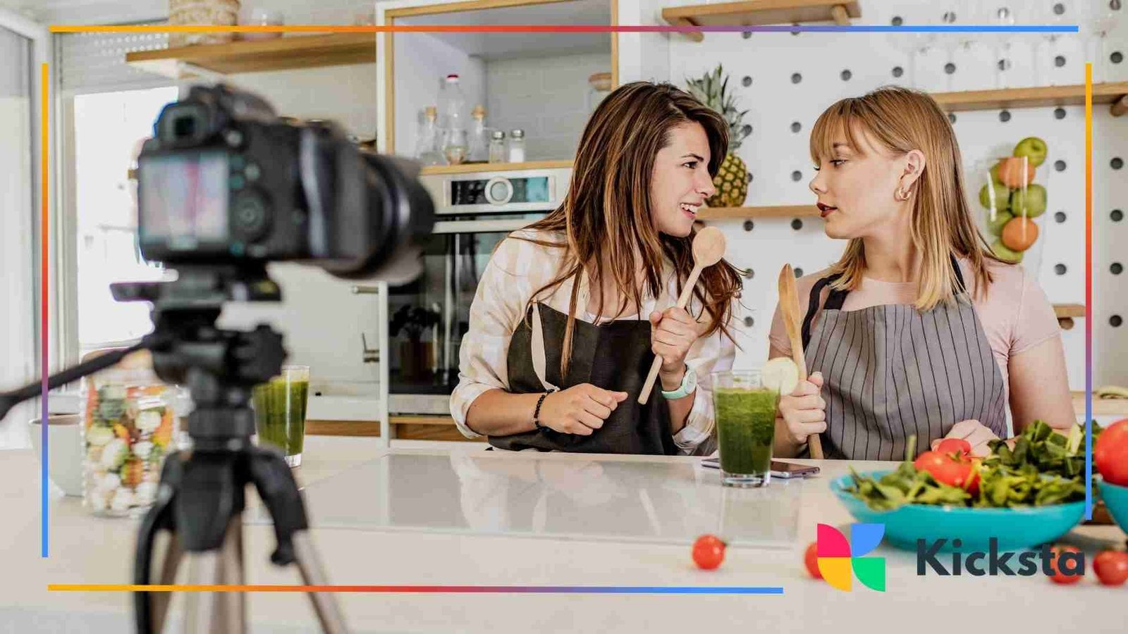 Two women in aprons filming a cooking video together in a kitchen, surrounded by fresh vegetables and a camera on a tripod.