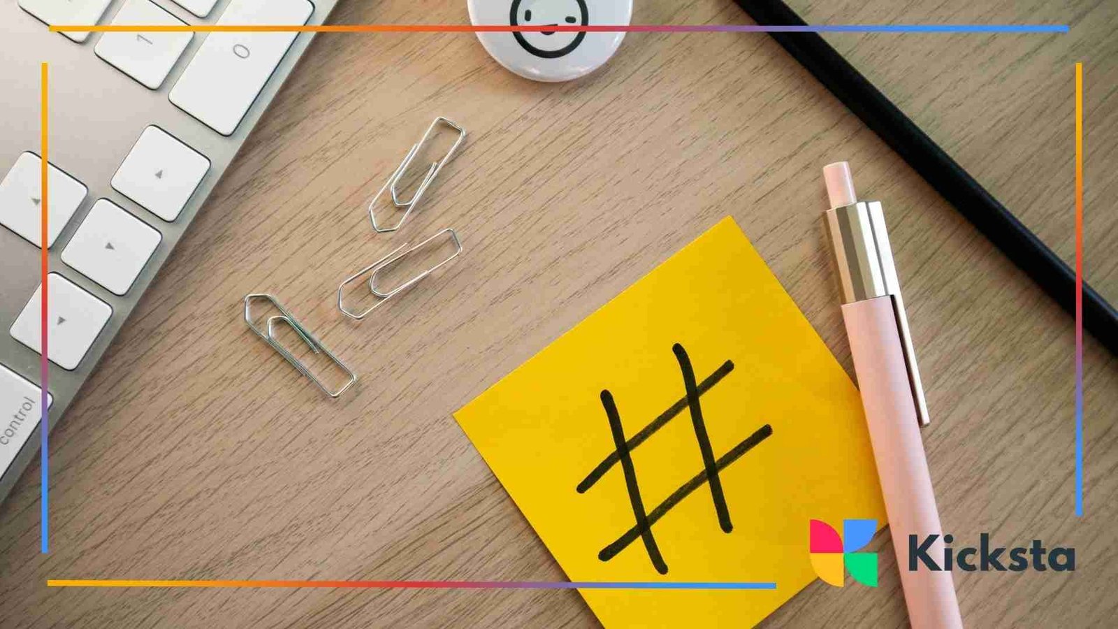 A yellow sticky note with a black hashtag drawn on it placed on a wooden desk beside a keyboard, paper clips, a pen, and a smiley-face button.