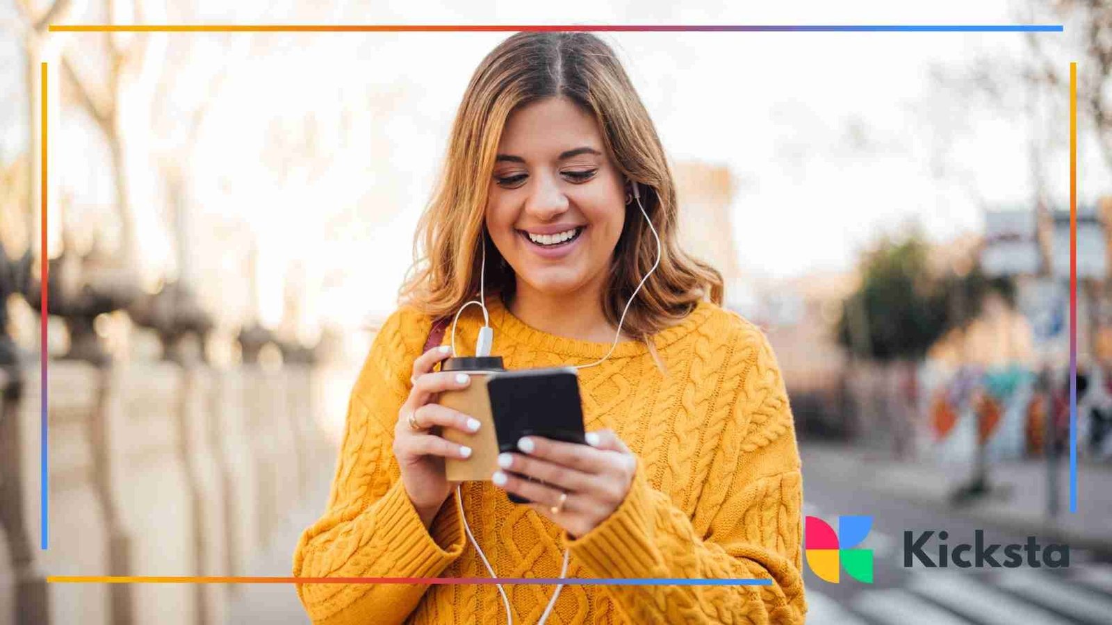 A woman in a yellow sweater smiles while looking at her phone and holding a coffee cup outdoors.