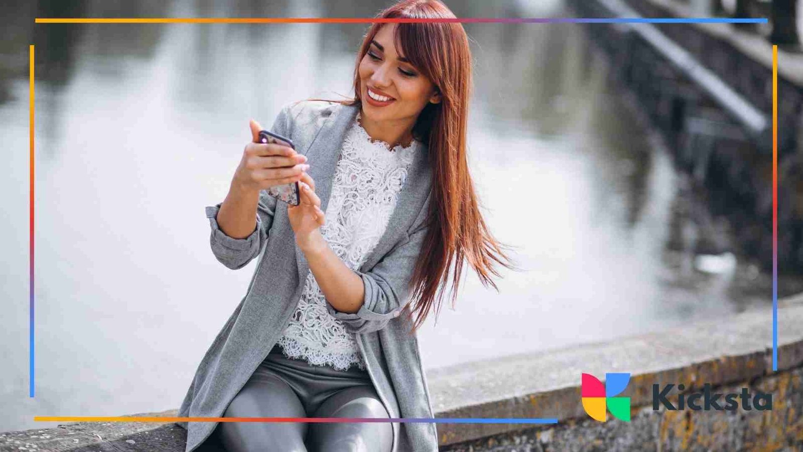 A woman sitting by a waterfront smiles while using her smartphone.