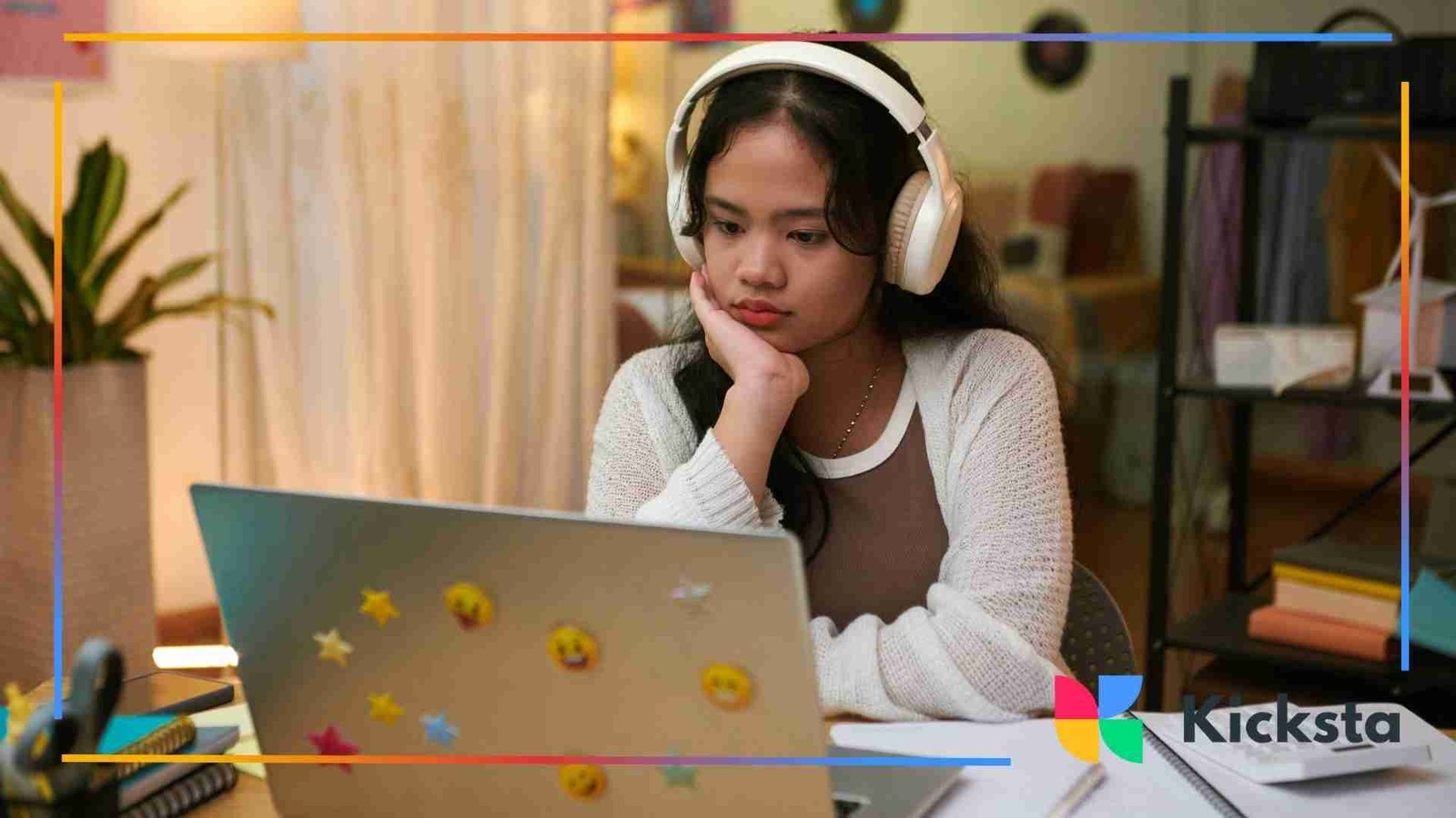 A young woman wearing large white headphones, resting her chin on her hand while looking thoughtfully at her laptop covered in emoji stickers.