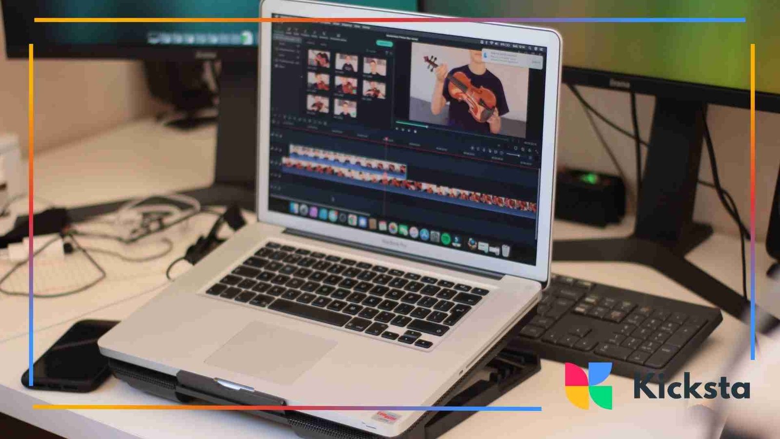 A laptop on a desk displaying a video editing program with clips of a person holding a violin, surrounded by a keyboard, monitor, phone, and cables.