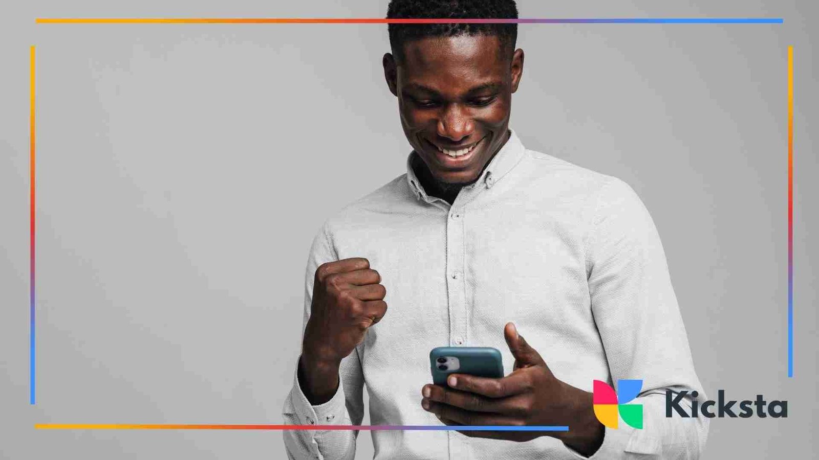 Man smiling and celebrating while looking at his phone, wearing a light gray shirt against a plain gray background.