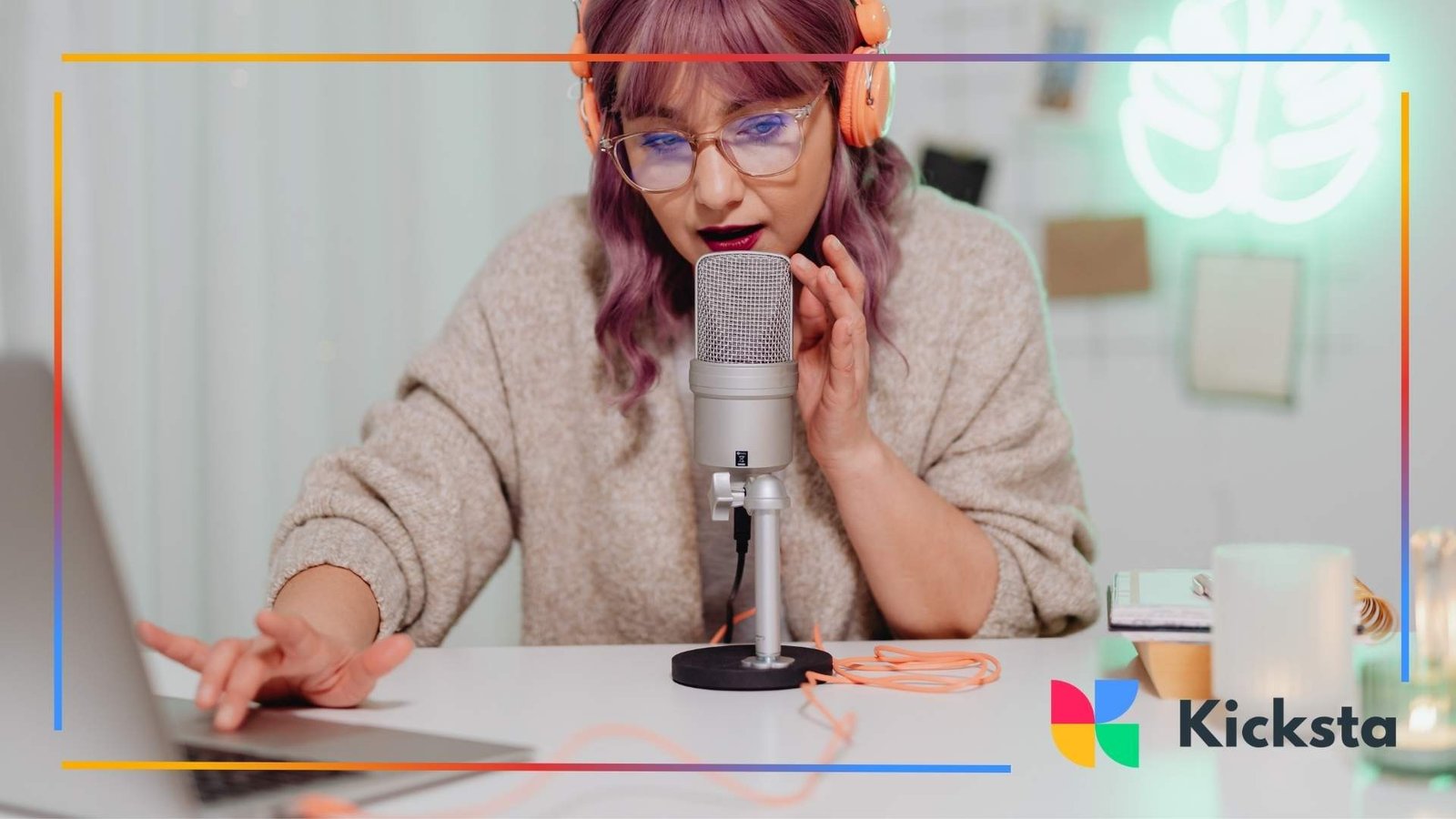 Young woman with purple hair wearing headphones and speaking into a microphone while working on her laptop.