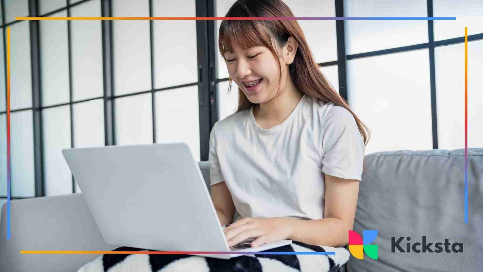 Young woman sitting on a couch working on a laptop, smiling while typing with a blanket over her legs.