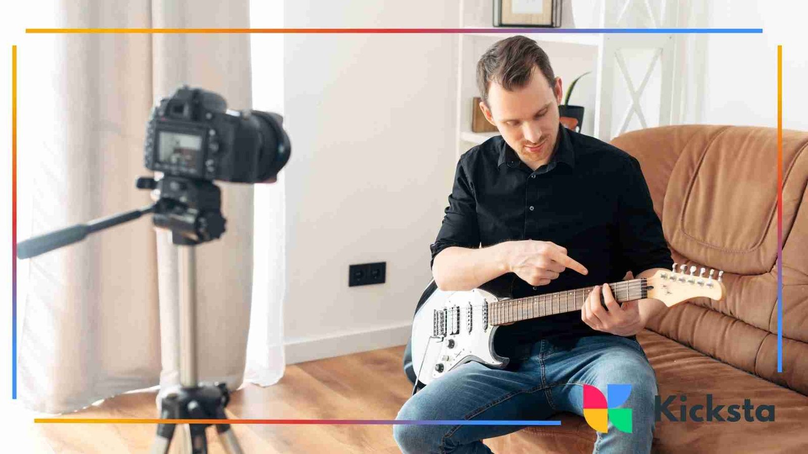 Man sitting on a couch recording a video tutorial while pointing at the fretboard of an electric guitar in front of a camera on a tripod.