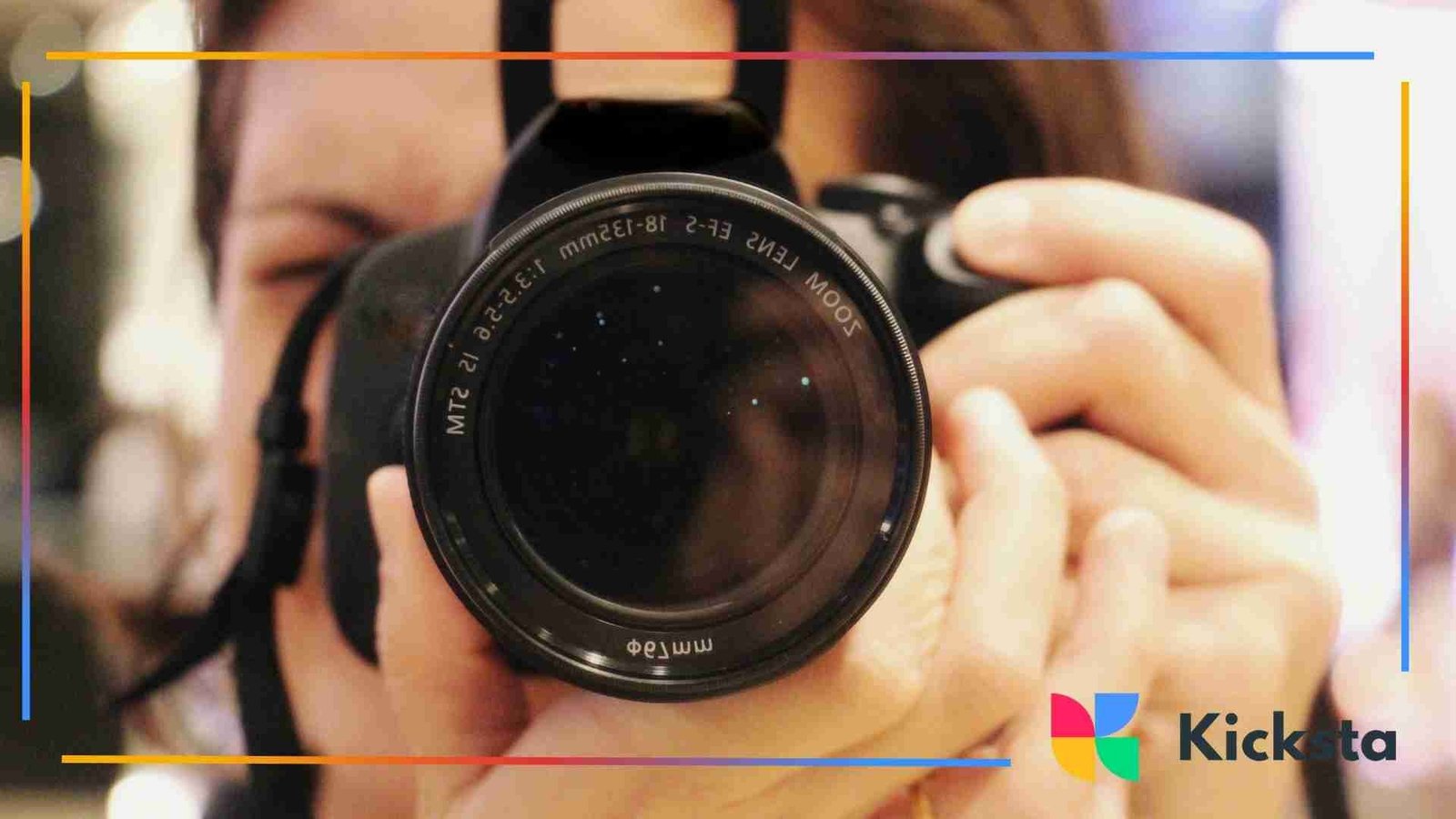 A person holding a DSLR camera directly toward the viewer, focusing through the lens in a blurred indoor setting.