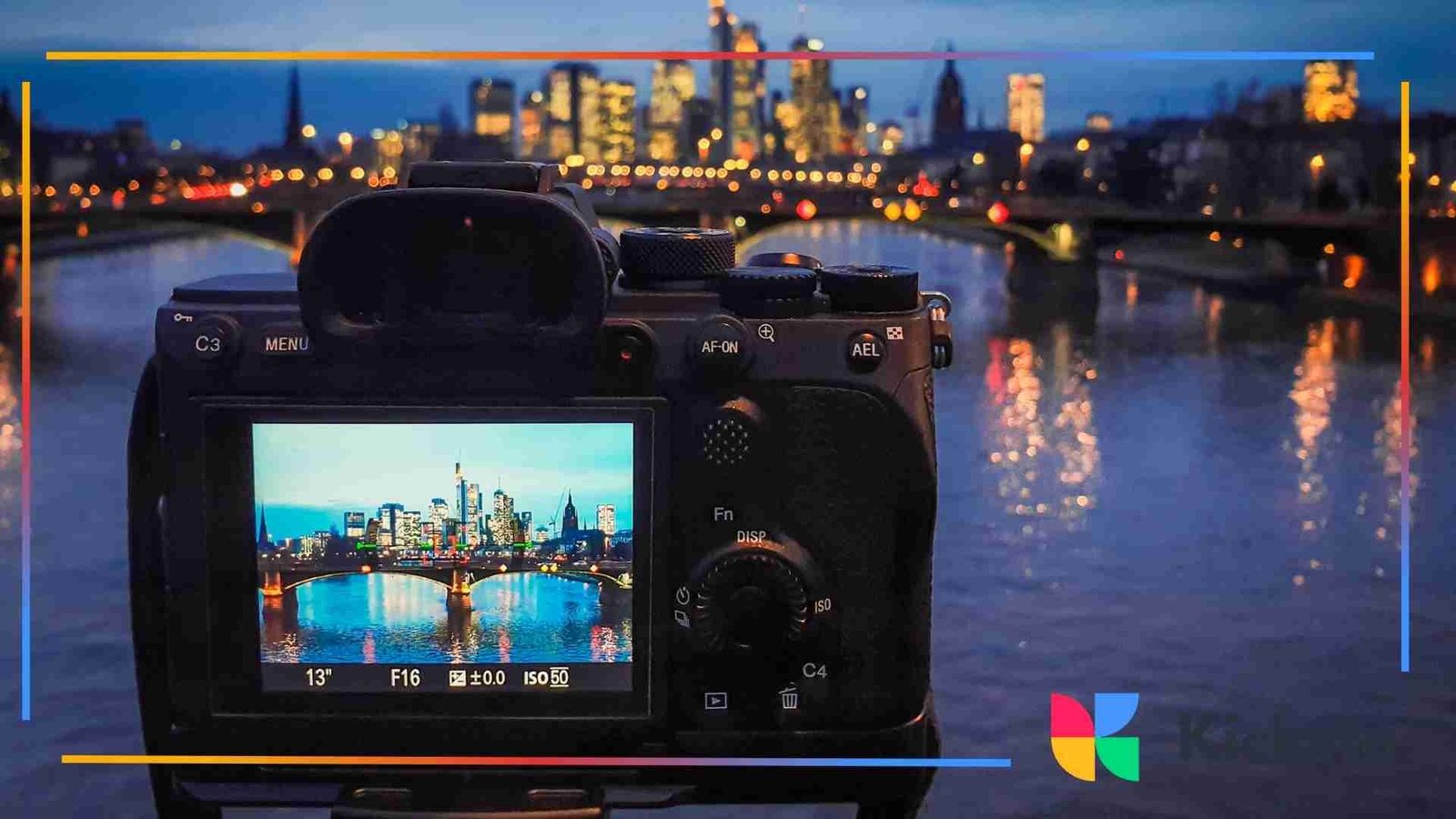 A camera on a tripod facing a river and illuminated city skyline at night, with the scene visible on the camera screen.