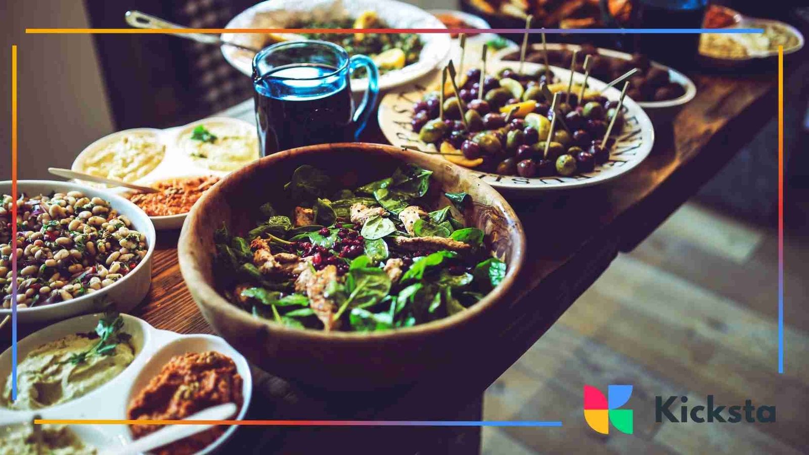Table filled with various dishes including a large wooden bowl of spinach salad, dips, olives, and small plates of colorful appetizers.