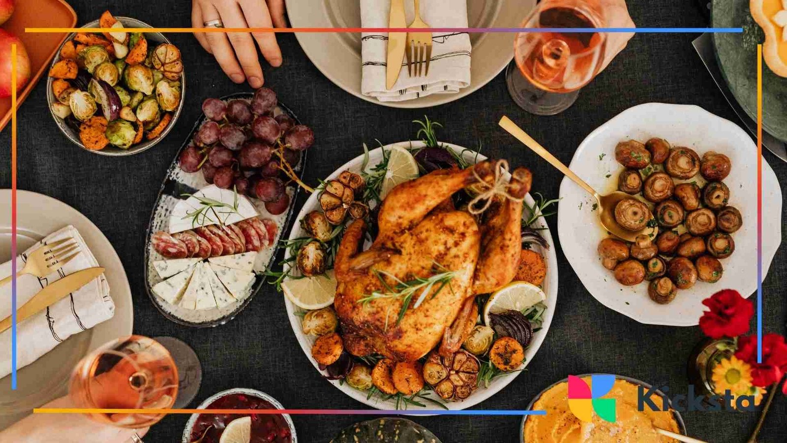 Overhead view of a large dinner table filled with roasted chicken, vegetables, cheese, grapes, and drinks, with people reaching for food.