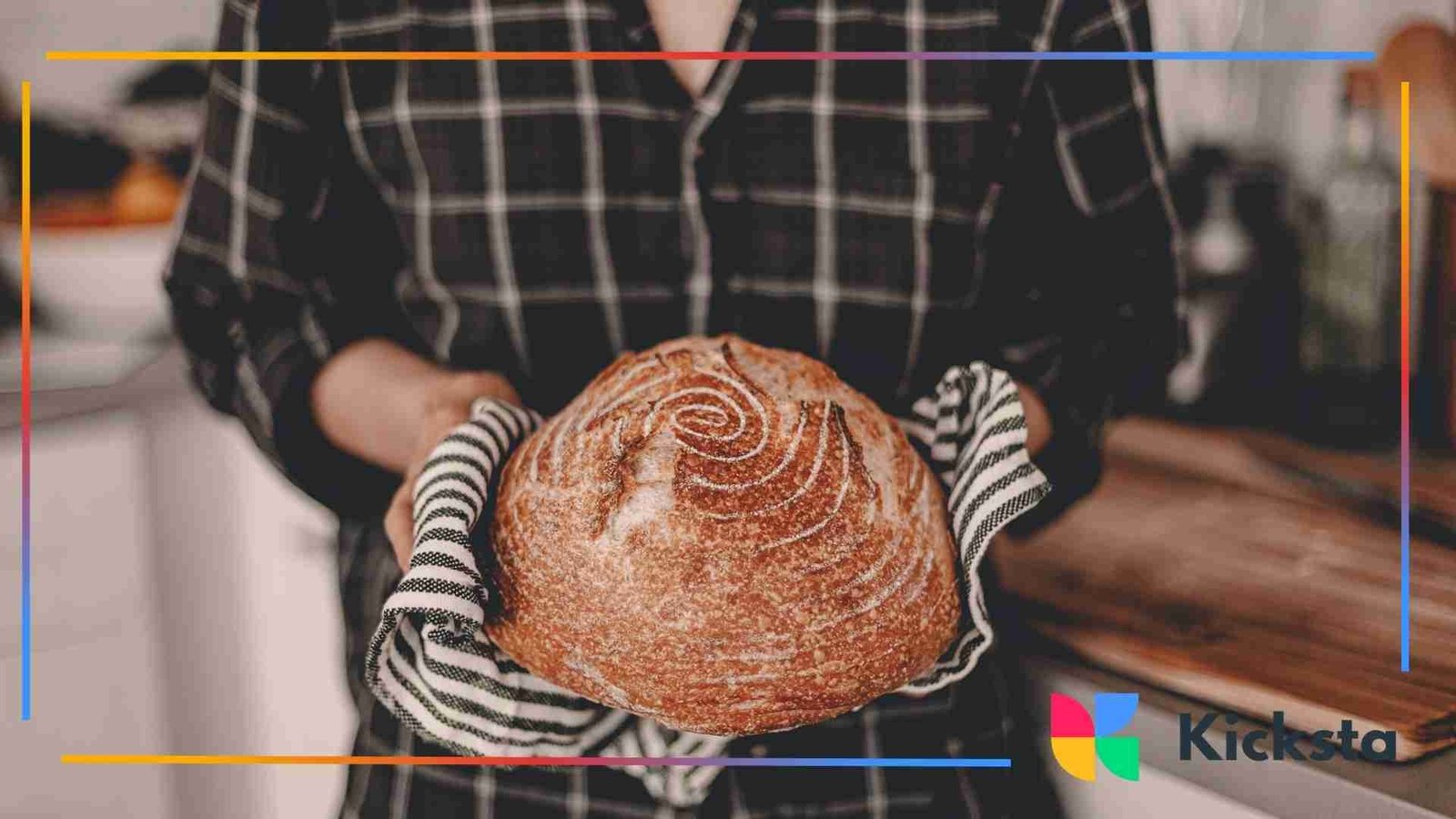 Person holding a freshly baked round loaf of artisan bread with a patterned crust, using striped oven mitts in a cozy kitchen.