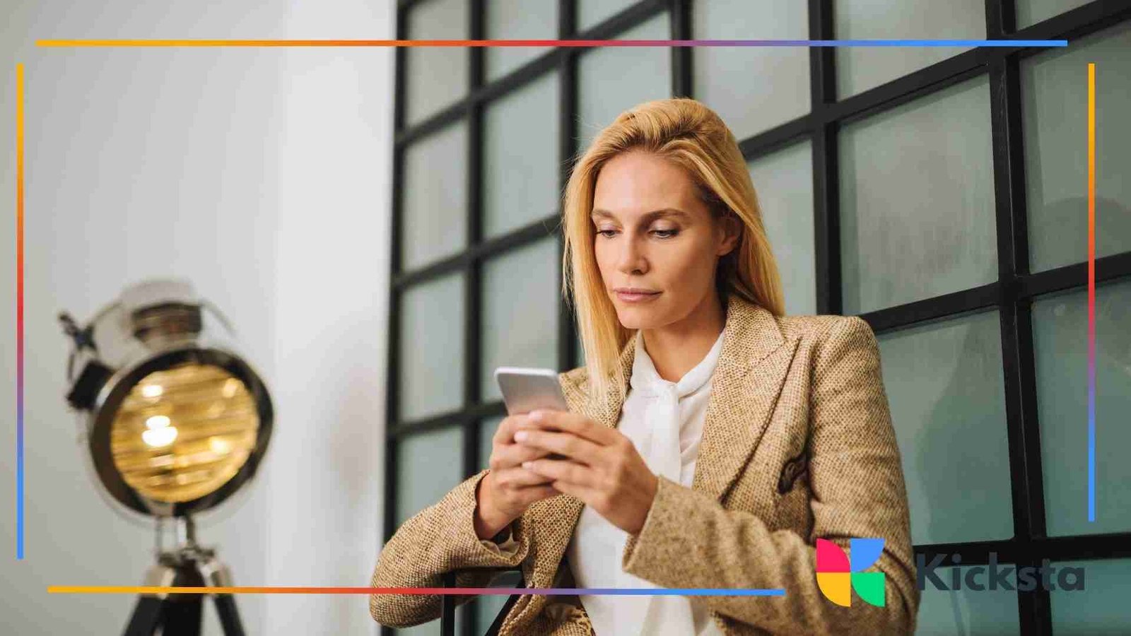 Woman wearing a tan blazer sitting indoors and focused on her smartphone.