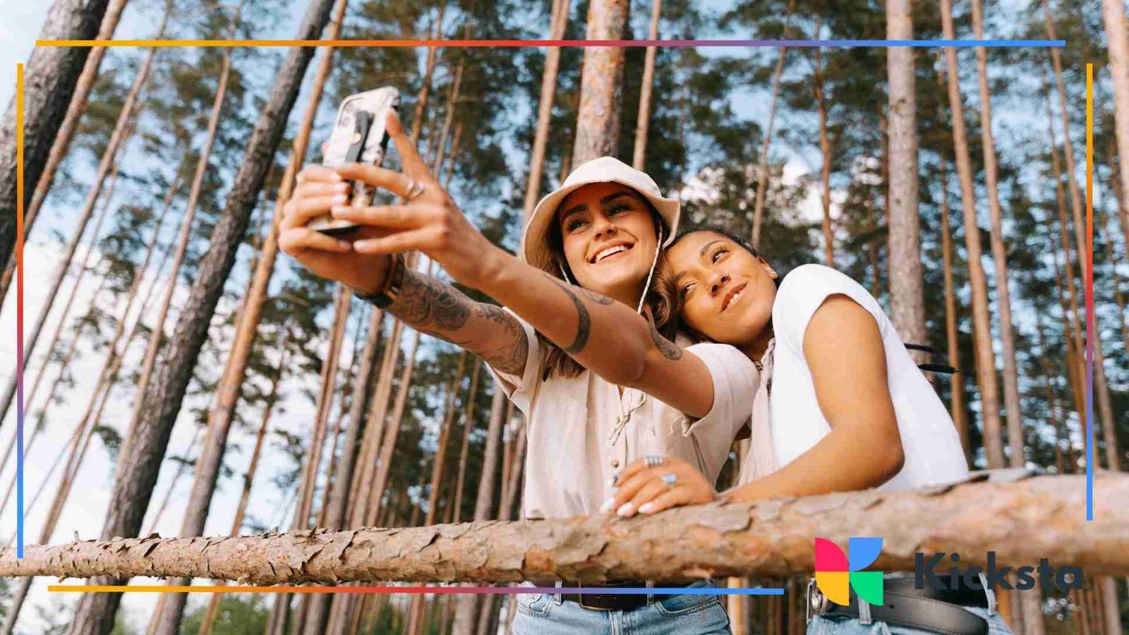 Two smiling friends taking a selfie together in a forest surrounded by tall trees.